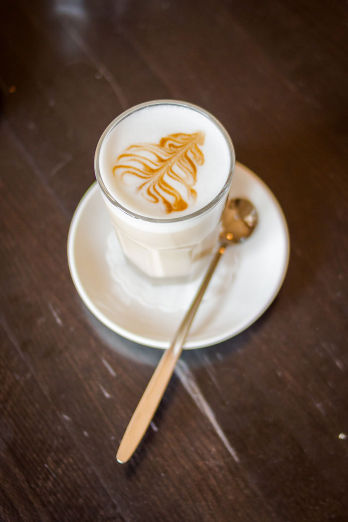 A latte with leaf-patterned foam art and a spoon on a saucer at A-Pine Restaurant Coffee Bar.